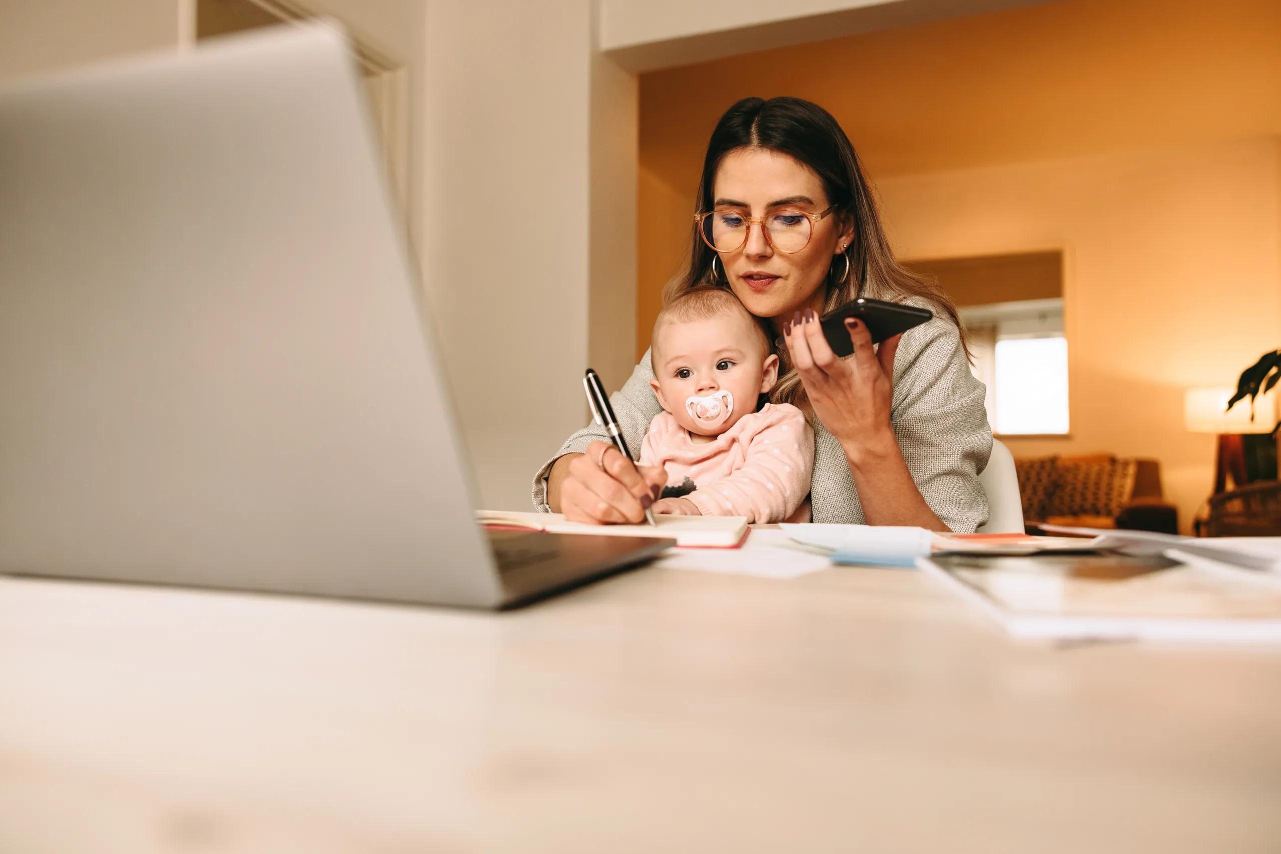 A mother working on her laptop with her baby in her arms