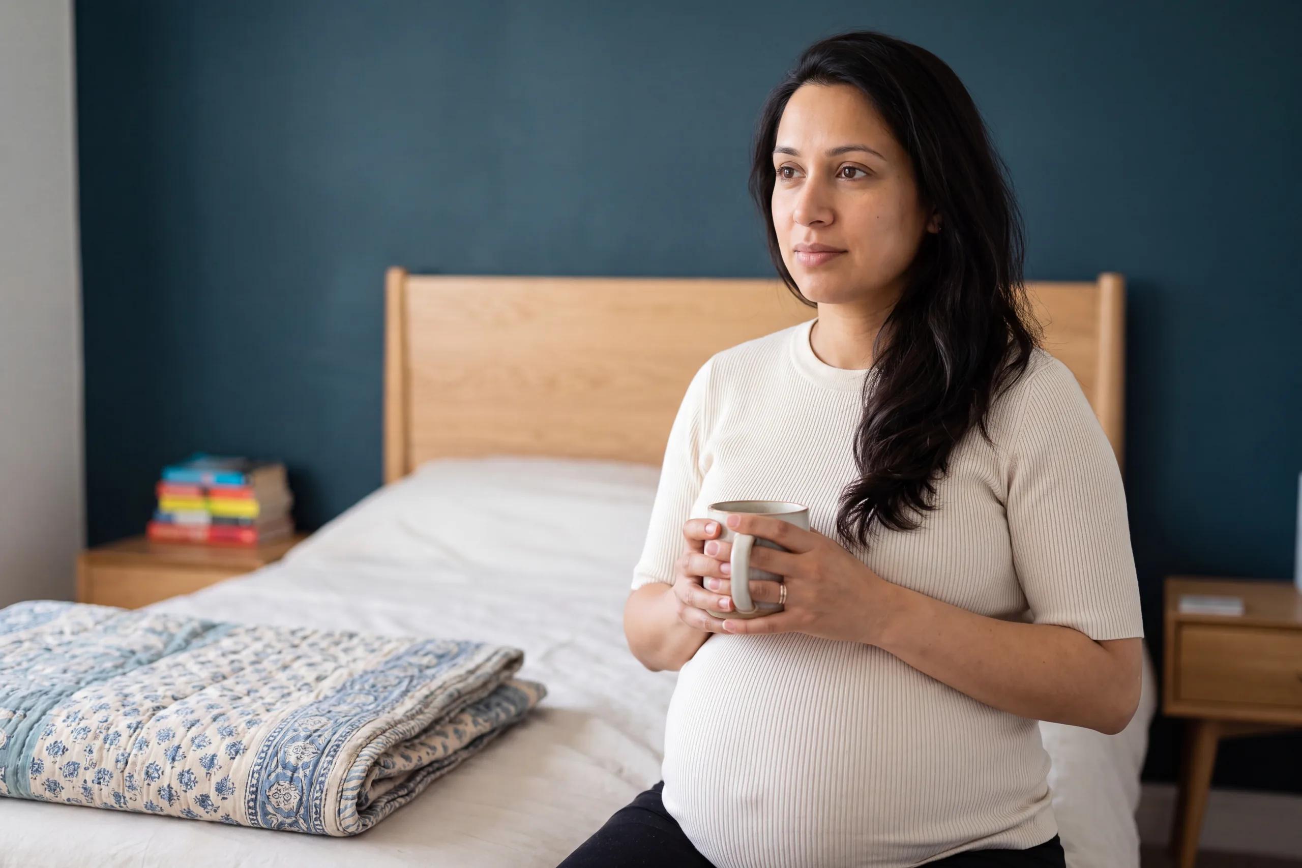Une femme enceinte assise au bord d’un lit, une tasse entre les mains.