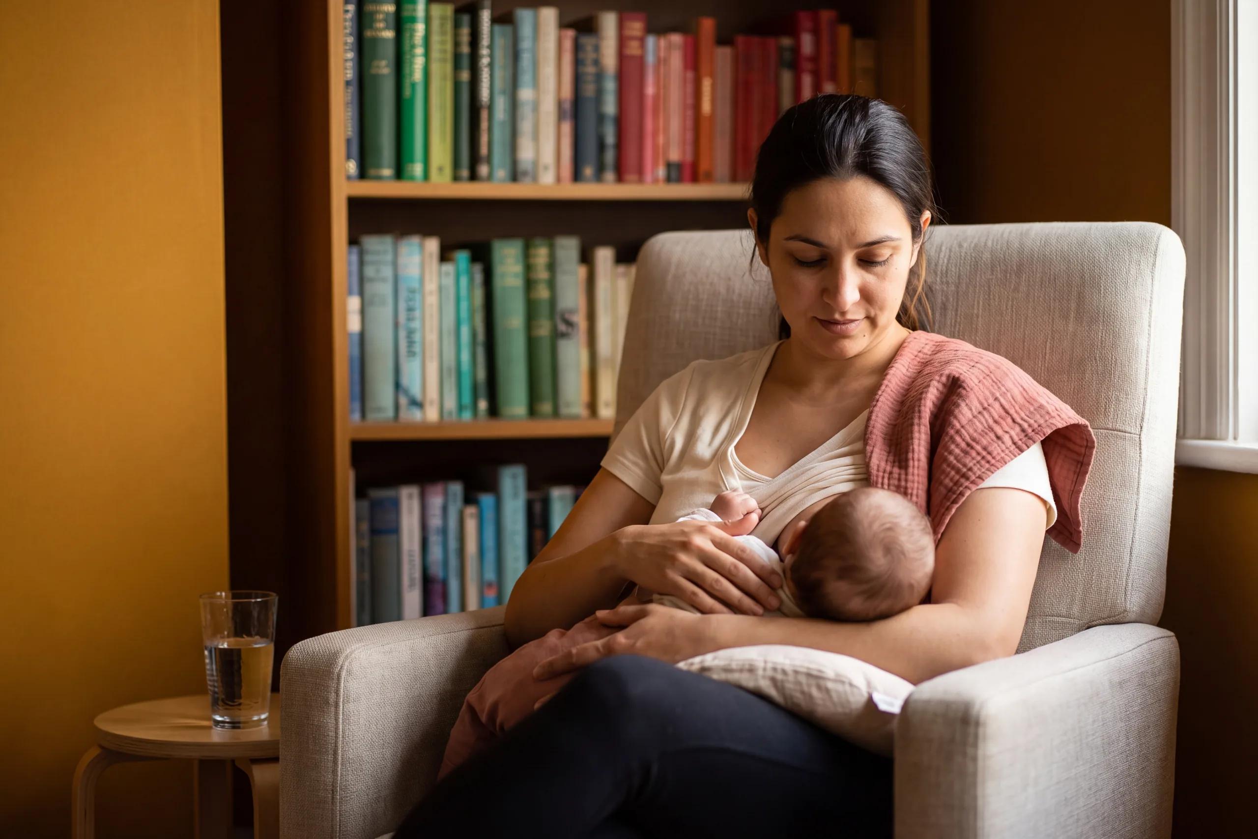 Une mère allaite son bébé dans un fauteuil, devant une bibliothèque.