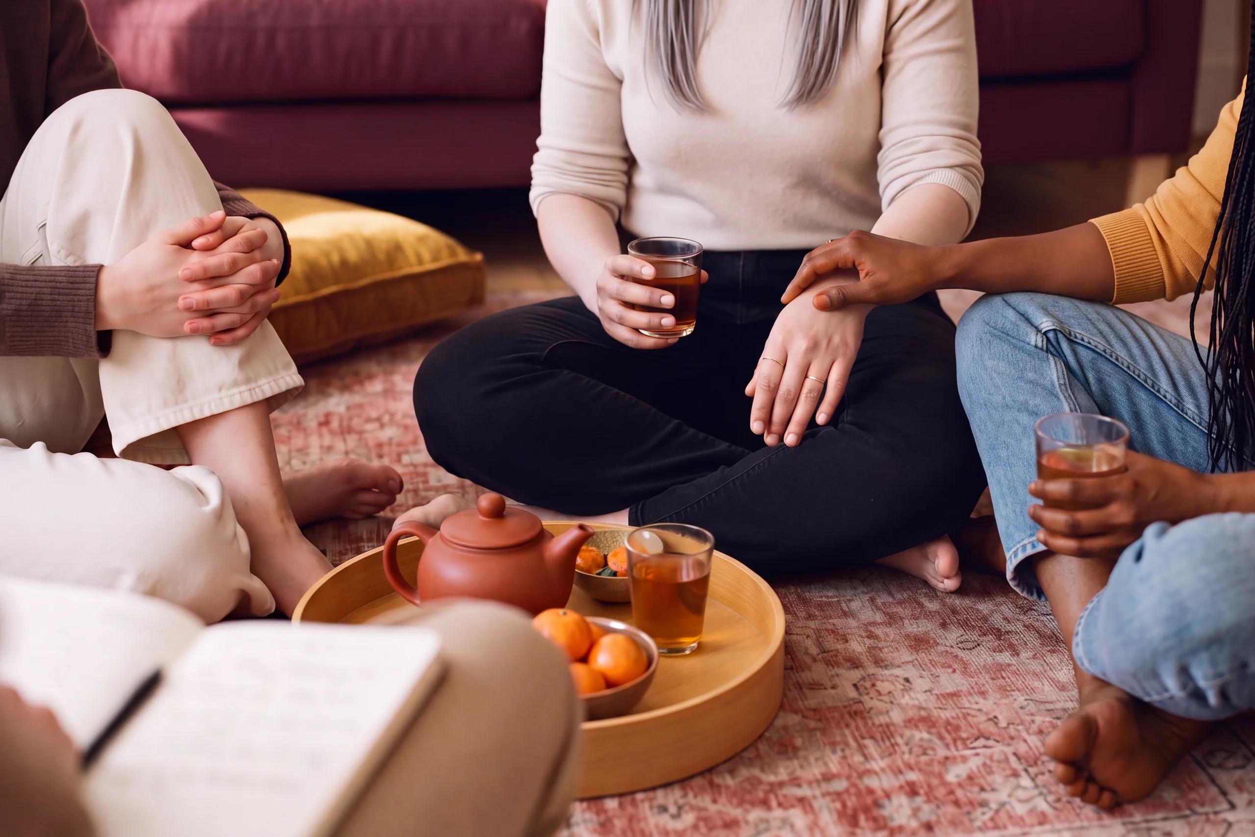 Quatre amies assises en cercle sur un tapis autour d’un plateau de thé ; l’une pose la main sur le bras de sa voisine